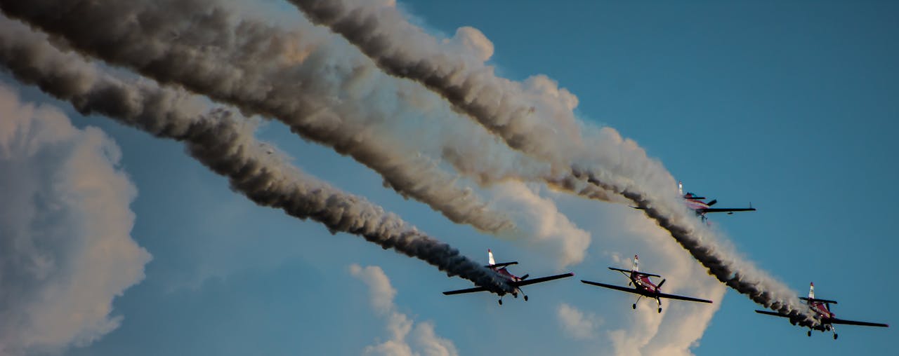 Four stunt airplanes perform dramatic aerobatic maneuvers leaving trails against a blue sky.